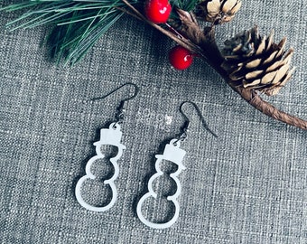 Op de afbeelding: Een paar witte sneeuwman-vormige oorbellen met zilveren haakjes. De oorbellen hangen aan een tak van een dennenboom met rode bessen.