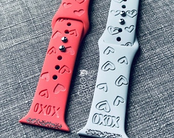 May include: Two Apple Watch bands with a heart pattern. The band on the left is pink with silver accents and the band on the right is light gray with silver accents. Both bands have the text "XOXO" printed on them.