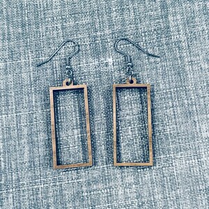 Op de afbeelding: Een paar houten oorbellen met een rechthoekig frame. De oorbellen zijn lichtbruin van kleur en hebben een zilveren haak.