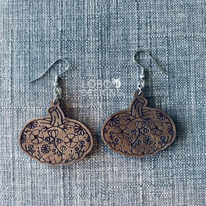 May include: A pair of wooden pumpkin earrings with a floral design. The earrings are brown and have silver hooks.