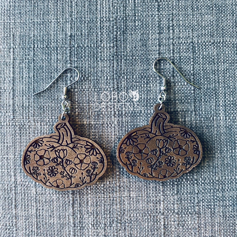 May include: A pair of wooden pumpkin earrings with a floral design. The earrings are brown and have silver hooks.