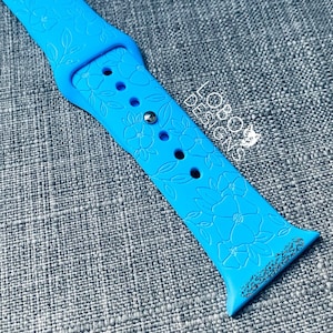 May include: A light blue silicone Apple Watch band with a floral design. The band has a silver buckle and is embossed with a floral pattern.