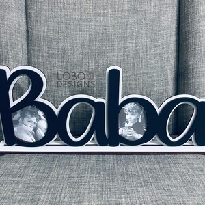 Puede incluir: Marco de fotos de madera negro con la palabra "Baba" escrita en letras blancas. El marco tiene dos aberturas para fotos. El marco está sobre una superficie gris.