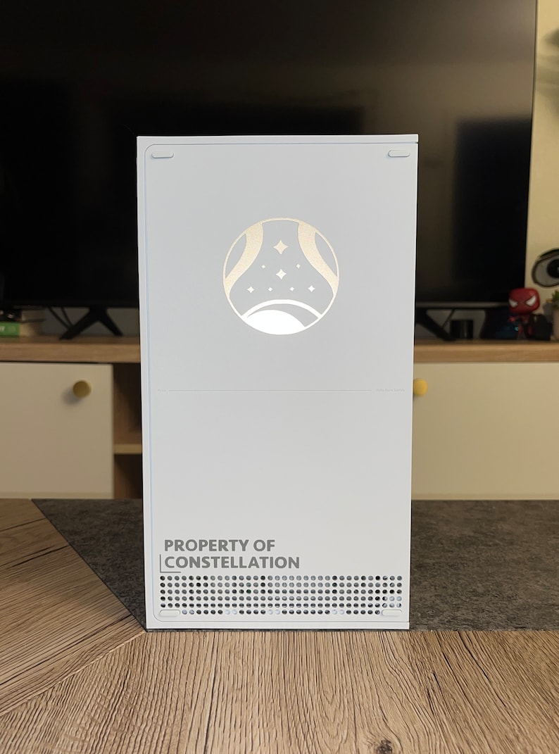 Op de afbeelding: Witte gameconsole met een zilveren logo van een halve maan en sterren. De console heeft een zwarte ventilatieopening en de tekst "PROPERTY OF CONSTELLATION" staat op de voorkant.