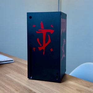 May include: A black gaming console with a red Doom logo on the front. The console is sitting on a wooden table.
