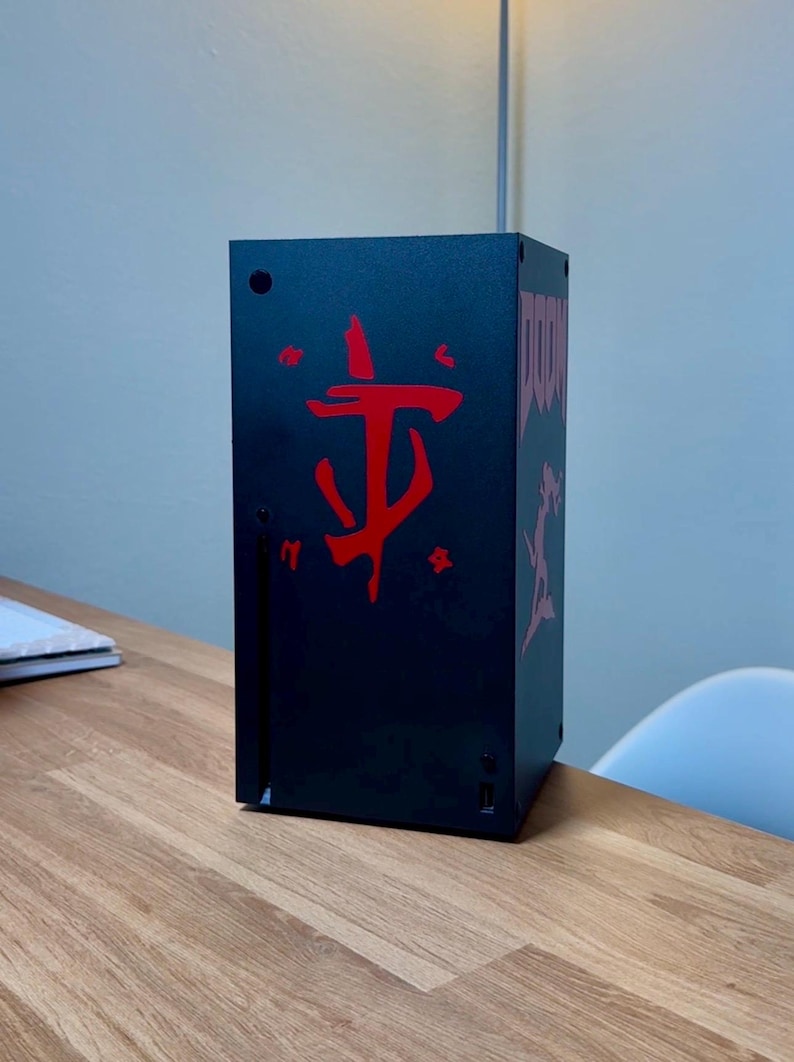 May include: A black gaming console with a red Doom logo on the front. The console is sitting on a wooden table.