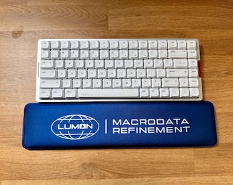 May include: A white keyboard with white keys and a silver frame sits above a blue wrist rest. The wrist rest has the text "LUMON | MACRODATA REFINEMENT" in white. The keyboard is on a wooden surface.