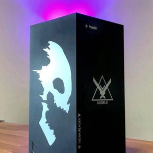 May include: A black gaming console with a white skull graphic on the front and a white triangle with a sword logo and the word "NOBLE" on the side. The console has a power button and a media reader.