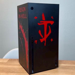 May include: A black gaming console with a red "REIGN IN HELL" text and a red Doom Slayer symbol on the front.