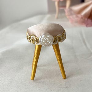 May include: Miniature gold-legged stool with a blush-colored, cushioned seat. The seat is trimmed with gold filigree and white rose accents. The stool's legs are painted gold, creating a delicate, decorative piece.