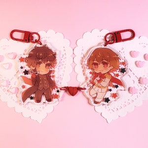 Peut inclure: Deux breloques de personnages avec des porte-clés rouges, représentant des illustrations de style anime de deux garçons. Les breloques sont reliées par une breloque en forme de cœur rouge. Les breloques sont sur un fond de dentelle blanche.