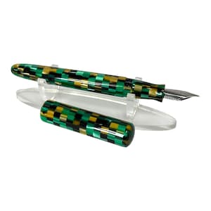 May include: A fountain pen with a checkerboard pattern in green, black, and gold. The pen is displayed with its cap off, revealing the nib. The pen rests on a clear acrylic stand against a white background.