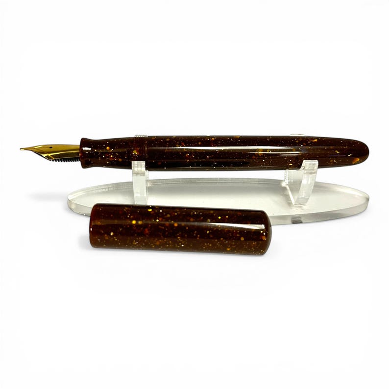 May include: A brown fountain pen with gold-colored nib and accents. The pen body and cap are speckled with gold flecks. The cap is detached and placed next to the pen on a clear acrylic stand.