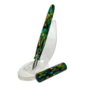 May include: A fountain pen with a checkerboard pattern in green, black, and gold. The pen is displayed on a clear plastic stand, with the cap removed and placed beside it. The nib is silver.