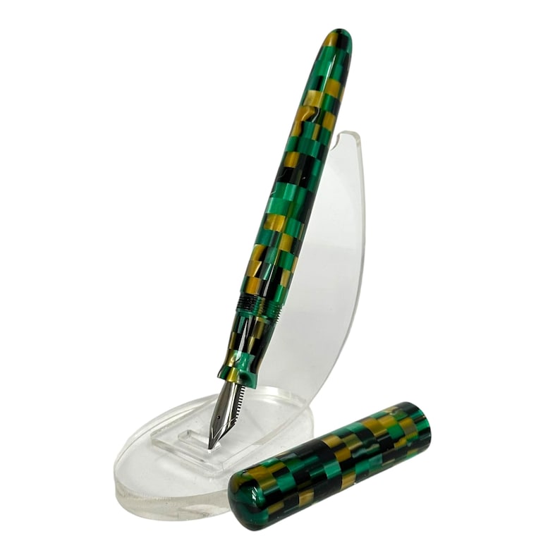 May include: A fountain pen with a checkerboard pattern in green, black, and gold. The pen is displayed on a clear plastic stand, with the cap removed and placed beside it. The nib is silver.