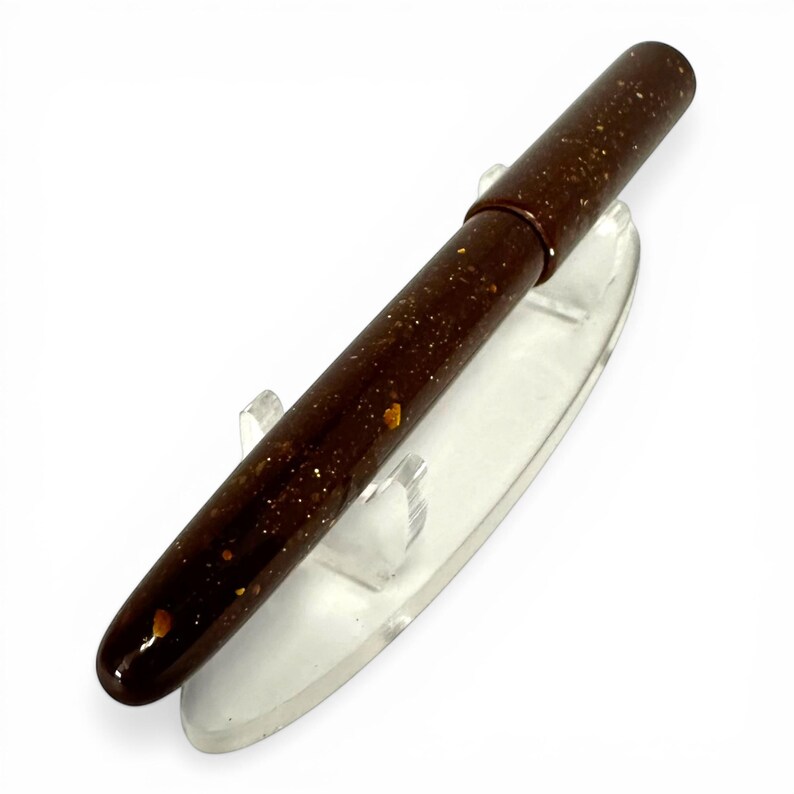 May include: A brown pen with gold flecks, resting on a clear plastic display stand. The pen has a rounded tip and a cap. The pen's body is a dark brown color with small gold-colored specks throughout.