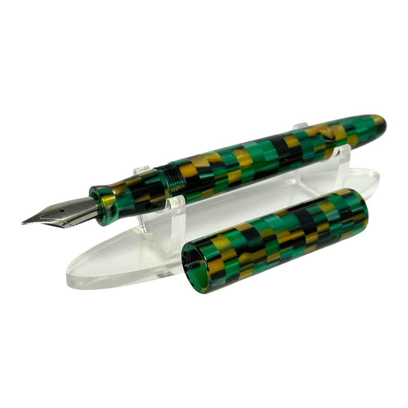 May include: A fountain pen with a checkerboard pattern in green, black, and gold. The pen has a silver nib and is displayed with its cap off on a clear stand. The pen's design is repeated on the cap.