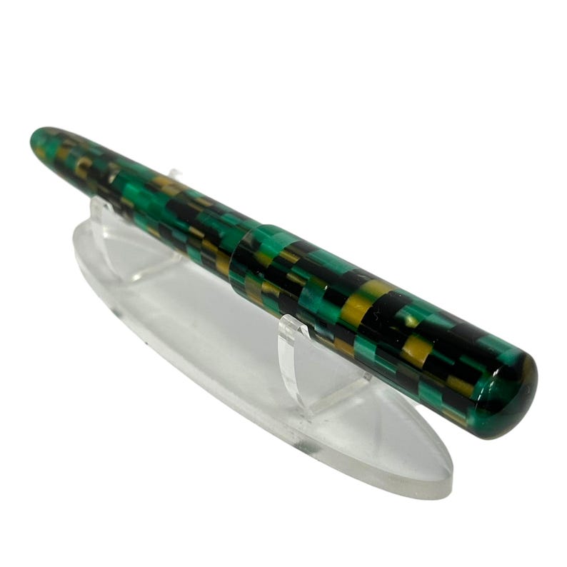 May include: A green, black, and gold patterned fountain pen rests on a clear acrylic stand. The pen's body features a mosaic-like design with a glossy finish. The pen is angled slightly, showcasing its full length and design.