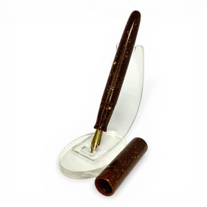 May include: A brown fountain pen with gold accents and a gold nib, displayed on a clear acrylic stand. The pen's body has a speckled design. The cap is detached and placed next to the pen.