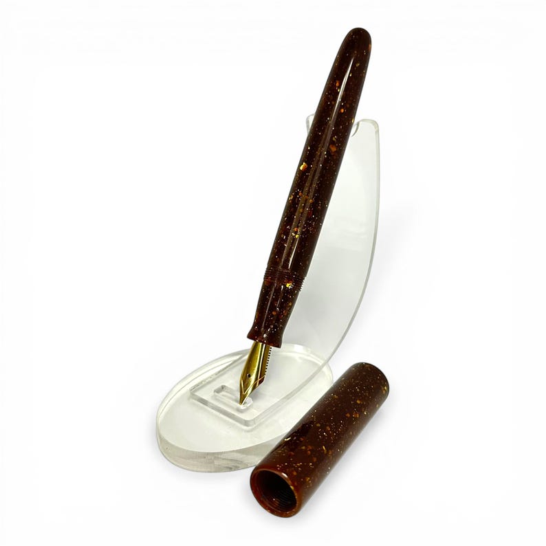 May include: A brown fountain pen with gold accents and a gold nib, displayed on a clear acrylic stand. The pen's body has a speckled design. The cap is detached and placed next to the pen.