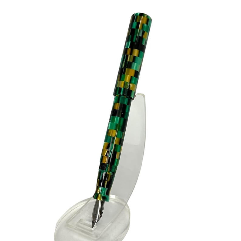 May include: A fountain pen with a checkered pattern in green, black, and gold. The pen has a silver nib and is displayed on a clear stand. The pen's design is a striking combination of colors and shapes.