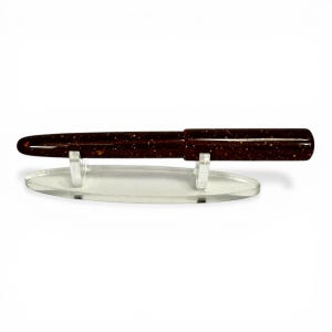 May include: A dark brown pen with gold flecks is displayed on a clear acrylic stand. The pen has a sleek, polished appearance. The stand is oval-shaped and has two small supports to hold the pen securely. The pen is a writing instrument.