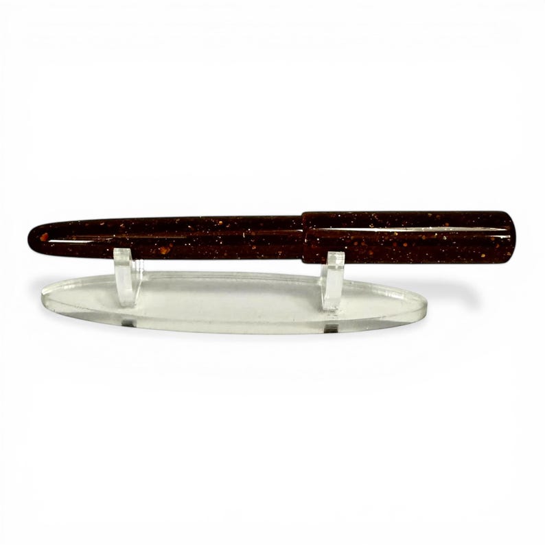 May include: A dark brown pen with gold flecks is displayed on a clear acrylic stand. The pen has a sleek, polished appearance. The stand is oval-shaped and has two small supports to hold the pen securely. The pen is a writing instrument.