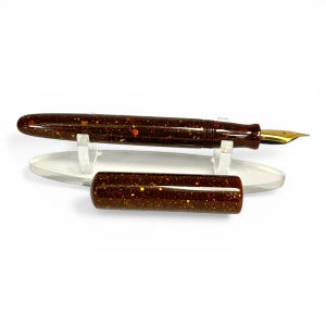 May include: A brown fountain pen with gold accents and a matching cap. The pen has a speckled design with gold and orange flecks. The pen is displayed on a clear acrylic stand against a white background.