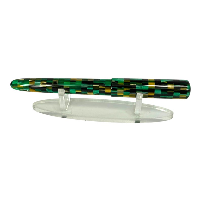 May include: A fountain pen with a green, black, and gold checkered pattern. The pen is displayed on a clear acrylic stand against a white background. The pen's design is a mix of rectangular shapes.