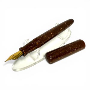 May include: A brown fountain pen with a gold nib and a matching cap. The pen has a speckled design and is displayed on a clear acrylic stand. The pen is a writing instrument.