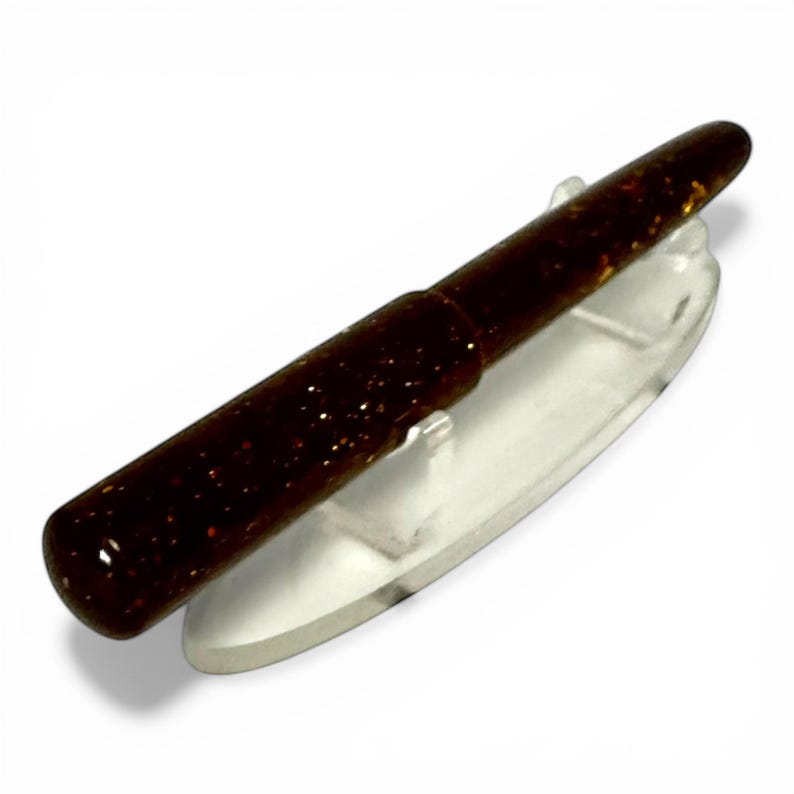 May include: A dark brown fishing lure with glittery flecks, resting on a clear plastic holder. The lure has a cylindrical body with a tapered end, designed for attracting fish. The background is white.