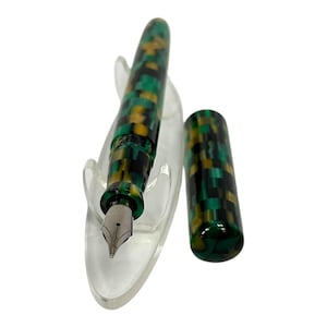 May include: A fountain pen with a green, black, and yellow mosaic pattern. The pen is displayed on a clear stand, with the cap removed and placed beside it. The nib is silver.
