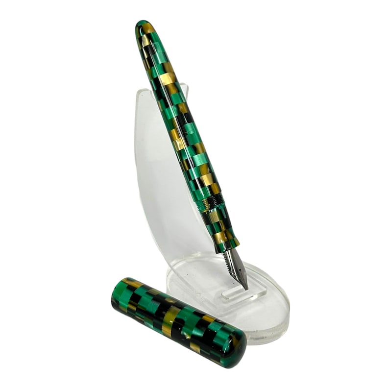 May include: A fountain pen with a checkerboard pattern in green, black, and gold. The pen is displayed on a clear acrylic stand, with the cap detached and placed beside it. The nib is silver.