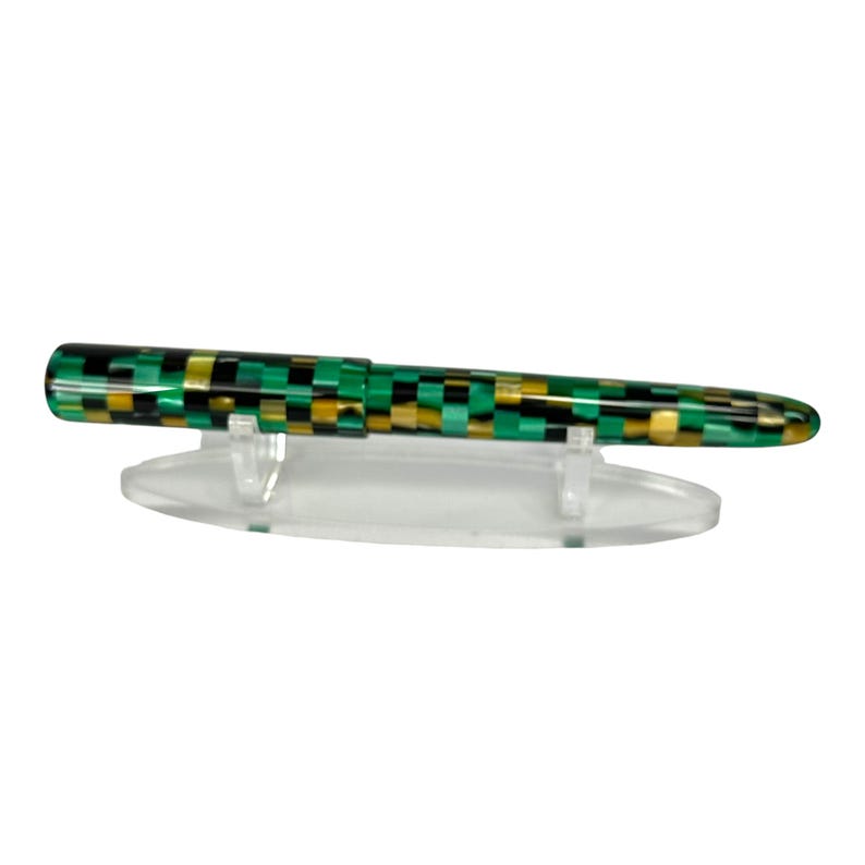 May include: A fountain pen with a checkered pattern of green, black, and gold. The pen is displayed on a clear plastic stand against a white background. The pen's design is a striking combination of colors.
