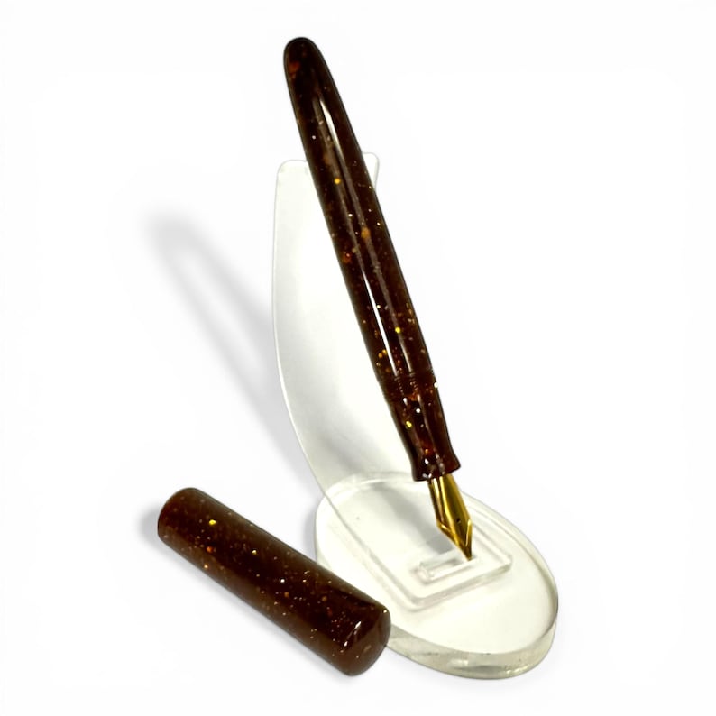 May include: A brown fountain pen with gold flecks and a gold nib, displayed on a clear acrylic stand. The pen's cap is placed separately. The pen is a writing instrument.