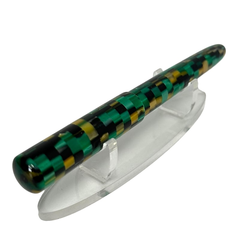 May include: A colorful fountain pen with a checkerboard pattern in green, black, and yellow. The pen is displayed on a clear acrylic stand, showcasing its unique design and craftsmanship. The pen's barrel has a glossy finish.
