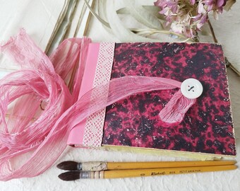May include: A pink and black marbled journal with a pink ribbon and a white button. The journal is surrounded by pink fabric and two paintbrushes.