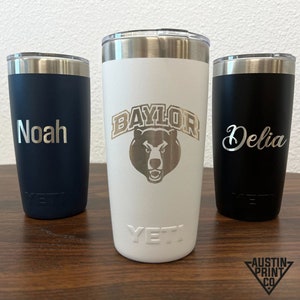 Puede incluir: Tres vasos Yeti personalizados. El vaso blanco tiene el logotipo de la Universidad Baylor y el texto "Baylor". El vaso azul marino tiene el nombre "Noah" grabado. El vaso negro tiene el nombre "Delia" grabado.