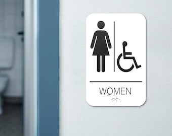 Può includere: Un cartello bianco con testo e simboli neri. Il cartello recita "DONNE" con una figura femminile e un simbolo di sedia a rotelle.