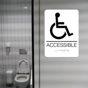 May include: White sign with a black wheelchair symbol and the word "ACCESSIBLE" in black text. Braille dots are below the word.
