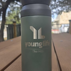 May include: A green insulated tumbler with a black lid. The tumbler has the Young Life logo and the text "The Hills" printed on it. The tumbler is sitting on a wooden surface.