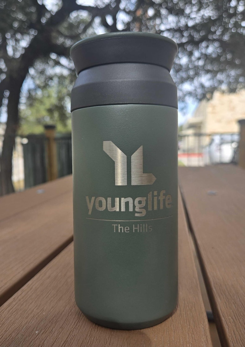 Pu&ograve; includere: Un bicchiere termico verde con coperchio nero. Il bicchiere ha il logo Young Life e la scritta "The Hills" stampati sopra. Il bicchiere &egrave; appoggiato su una superficie di legno.