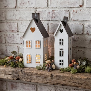 May include: Two white wooden house-shaped candle holders with dark grey roofs. Each has window cutouts and a heart-shaped opening. The candle holders are on a wooden shelf with greenery and dried flowers.