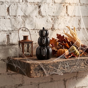 May include: A black ceramic stacked pumpkin figurine with the word BOO printed vertically in white letters, positioned next to a rustic metal lantern and a cluster of dried autumn leaves and small gourds on a weathered wooden shelf.