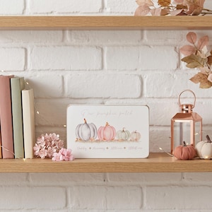 May include: A rectangular sign with the text "Our pumpkin patch" and illustrations of pumpkins in shades of grey, pink, and green. The sign is on a wooden shelf with books, flowers, and a lantern. The text "Daddy, Mummy, Little one, Littlest one" is below the pumpkins.