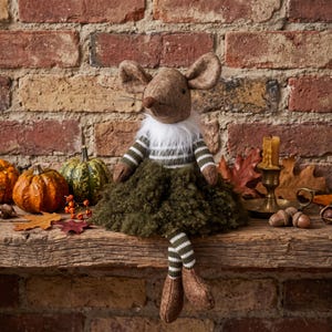 May include: A plush mouse figurine with large ears, a brown fabric head, and a white faux fur collar. It wears a green and white striped long-sleeve shirt, a dark green textured skirt, and matching striped leggings with brown fabric shoes.