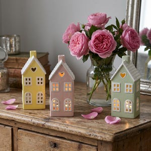 May include: Three ceramic house-shaped candle holders in yellow, pink, and green, each with a white roof and heart-shaped cutout. The candle holders are on a wooden surface with a vase of pink roses.