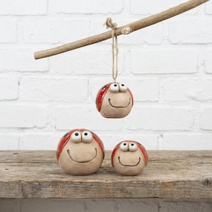 May include: Three ceramic ladybug ornaments with smiling faces. One is suspended from a wooden branch. The ladybugs have red shells with black spots and tan faces. They are arranged on a wooden surface.