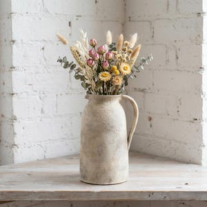 May include: A cream-coloured ceramic jug with a handle, holding a display of dried flowers. The bouquet features pink roses, yellow daisies, and different types of grasses. The jug is on a wooden surface against a white brick wall.