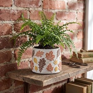 May include: A white ceramic planter with a textured surface featuring raised, colourful autumn leaf motifs in shades of orange, brown, and yellow. A vibrant green fern plant is potted inside the container, which has a cylindrical shape.